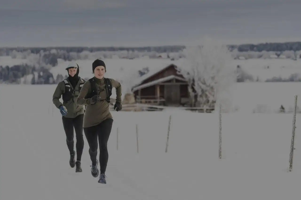 Två personer springer i snölandskap. De är klädda för att springa i kyla, med mössor, handskar och varma kläder. I bakgrunden syns ett snötäckt hus och träd.