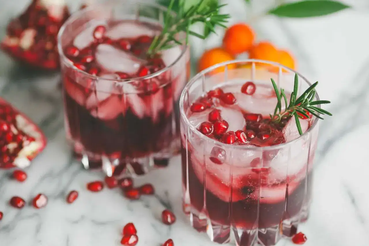 Orzeźwiający drink z sokiem z granatu, lodem i gałązką rozmarynu. Na stole rozsypane nasionka granatu.