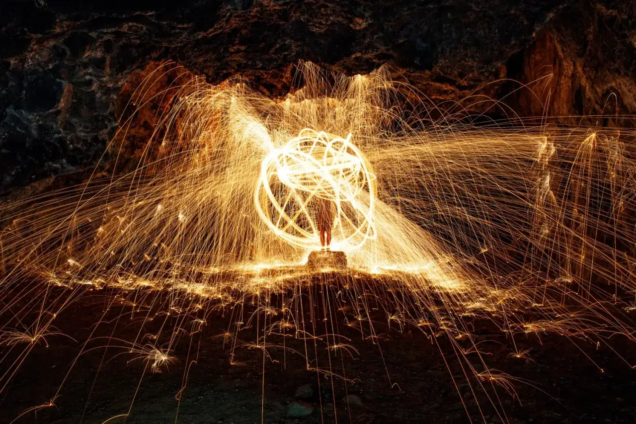 Una persona sta al centro di una grotta, circondata da un vortice di scintille dorate. L'arte di fotografare il fuoco crea un'aura luminosa e dinamica.