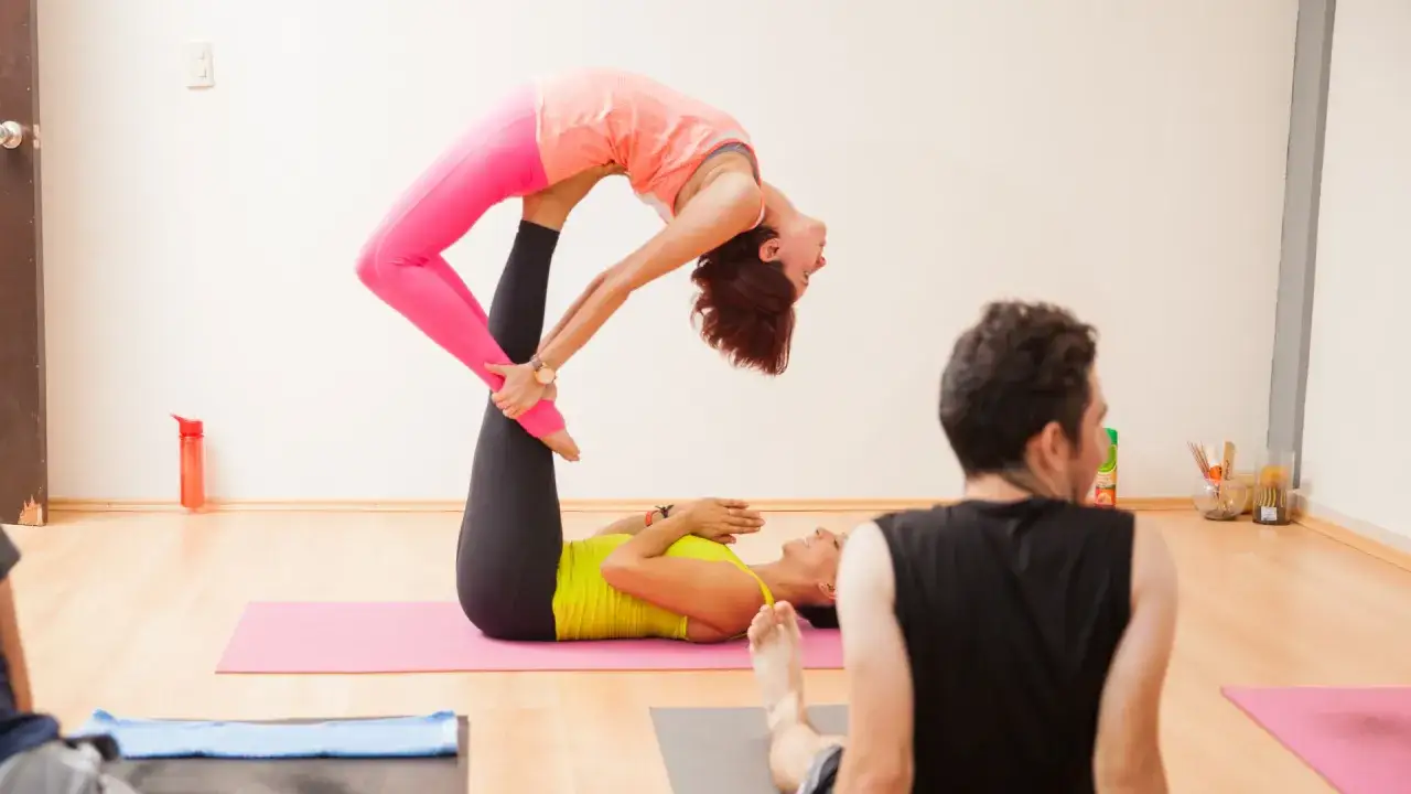 Dwie kobiety ćwiczą **acroyoga**. Jedna leży na macie, druga jest w powietrzu, wsparta na jej stopach.