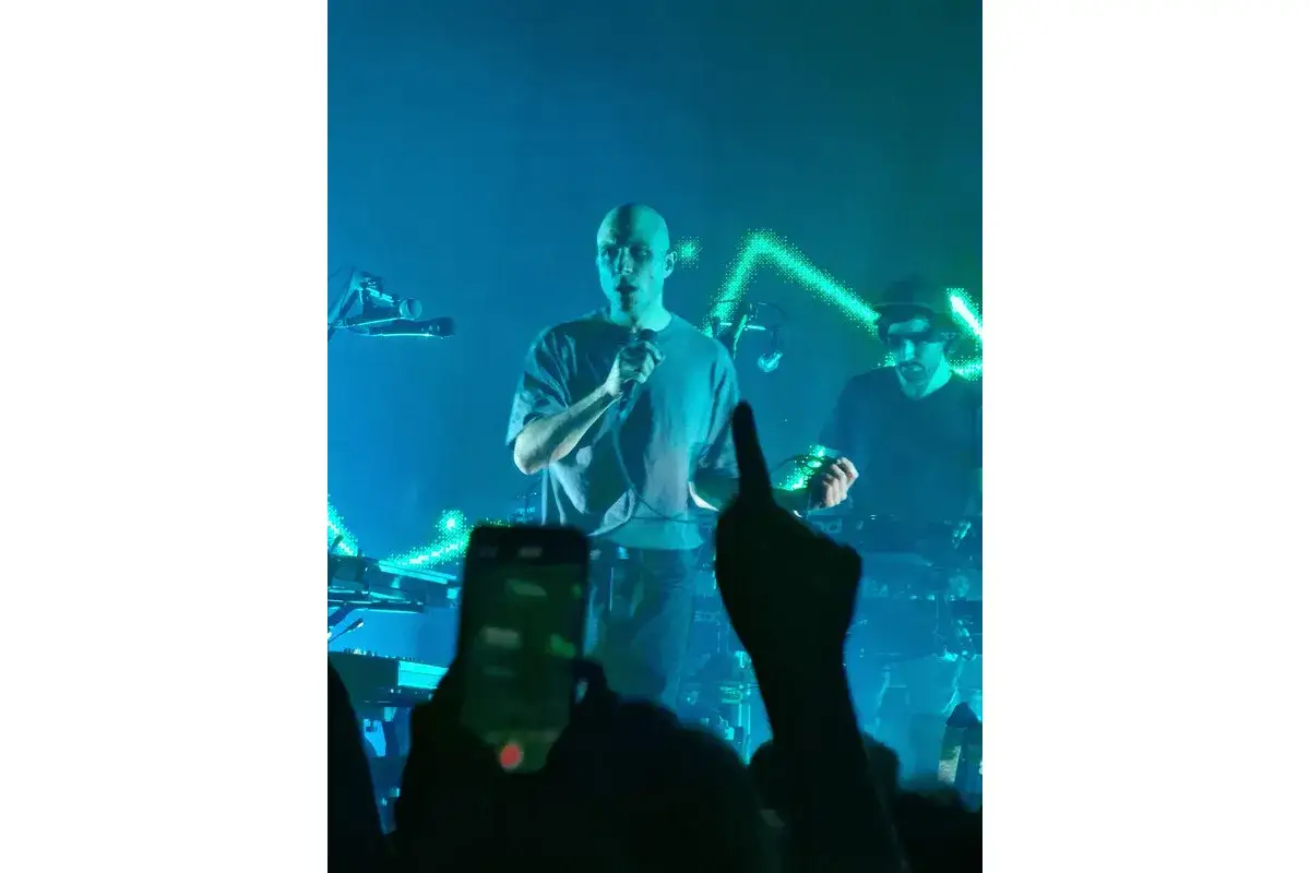 Sul palco, un uomo calvo canta al microfono, mentre un altro musicista suona dietro di lui. La scena è illuminata da luci blu e verdi, creando un'atmosfera da concerto. Sembra che la contessa e i cani stiano assistendo allo spettacolo.