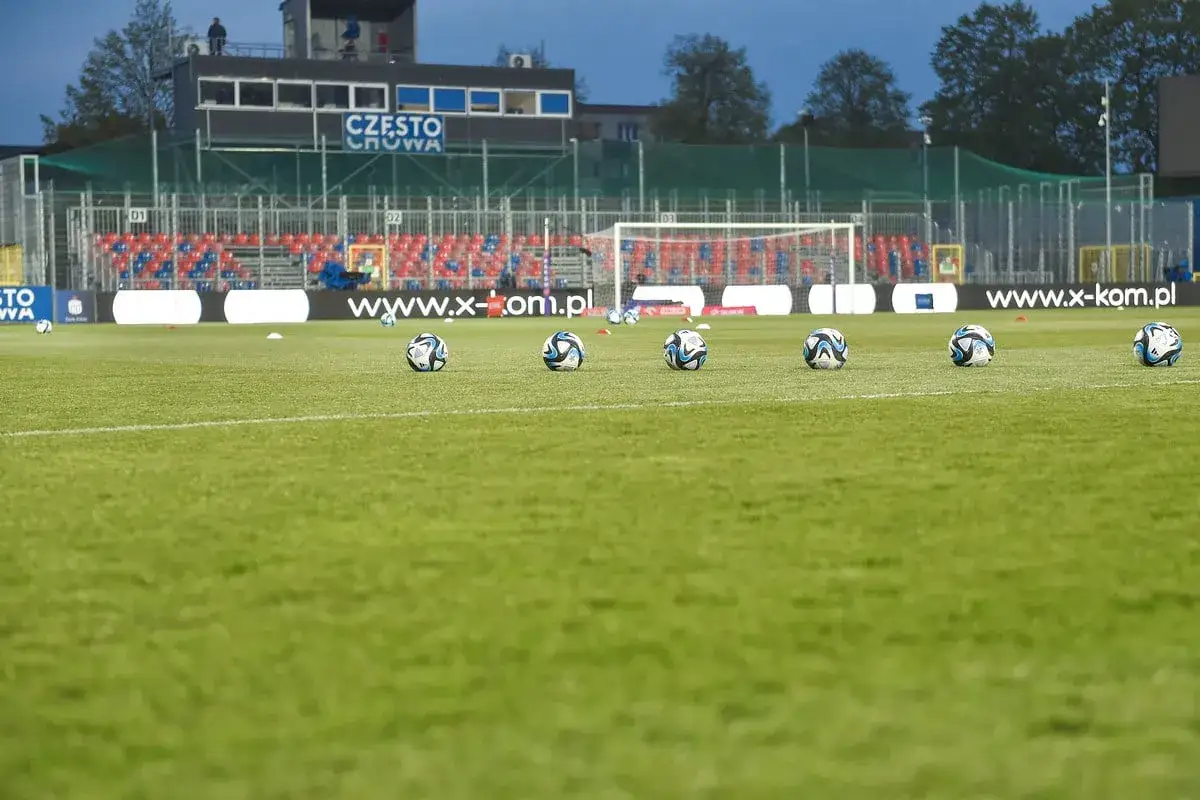 Piłkarze z Częstochowy szykują się do treningu. Na murawie stadionu leży kilka piłek, a w tle widać trybuny i bramkę.