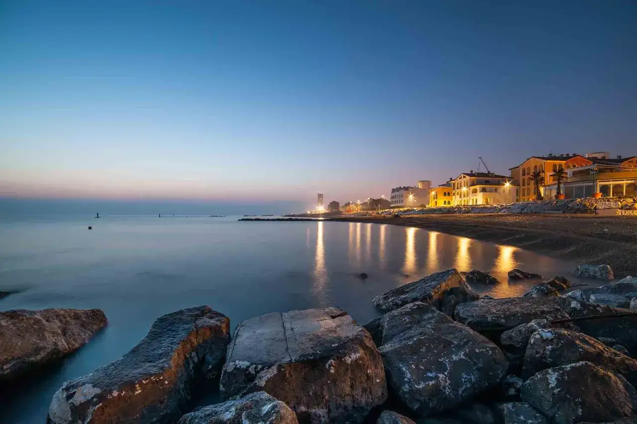 La sera a Porto Recanati, con le luci dei palazzi che si riflettono sul mare calmo e le rocce in primo piano.