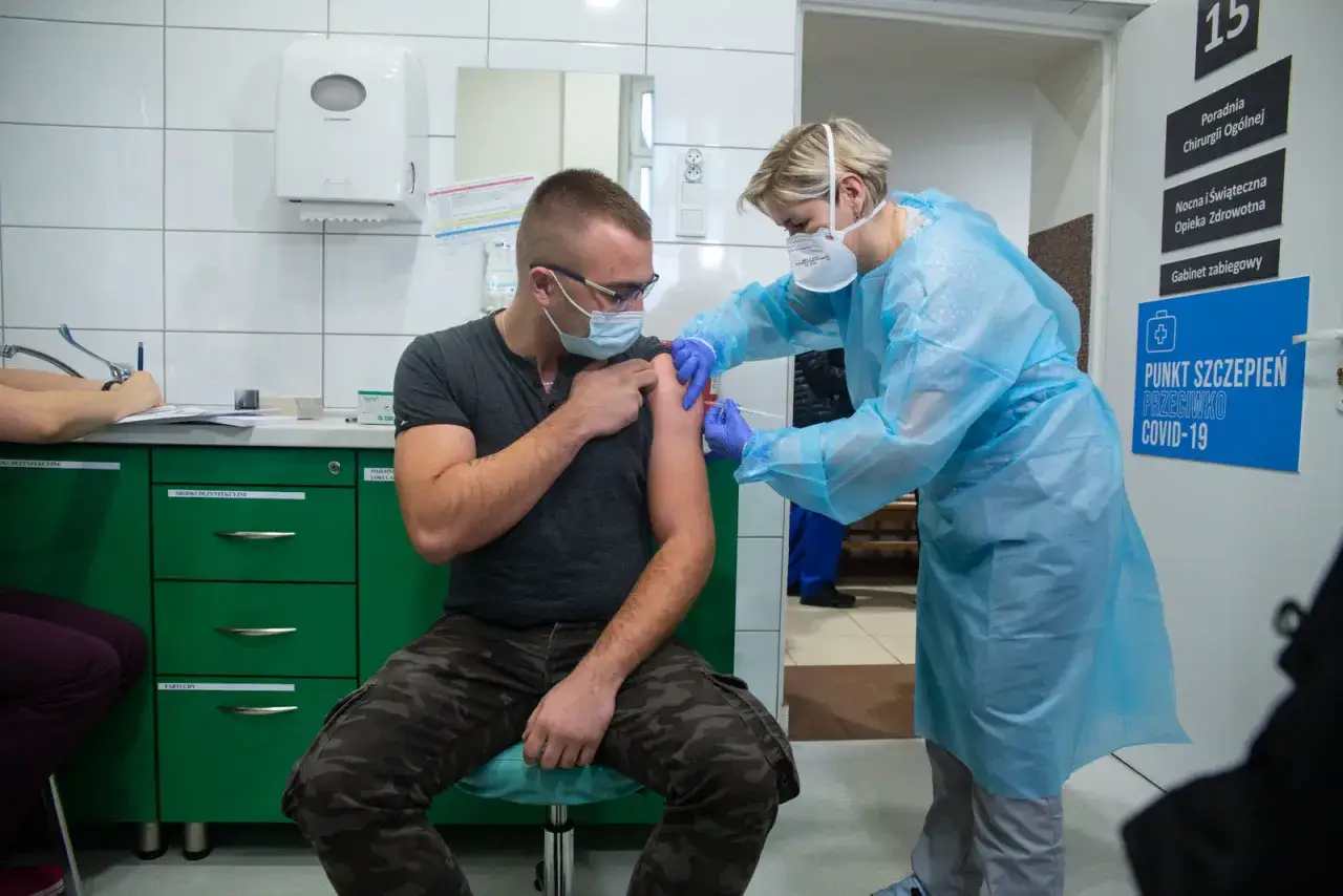 Mężczyzna otrzymuje szczepionki na covid w punkcie szczepień. Pielęgniarka w fartuchu zakłada mu zastrzyk.