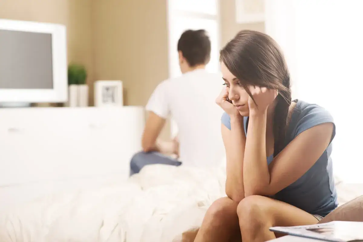 Mujer pensativa en la cama, con su pareja de espaldas. Ella asume su rol de salvadora, esperando una reconciliaci&oacute;n.