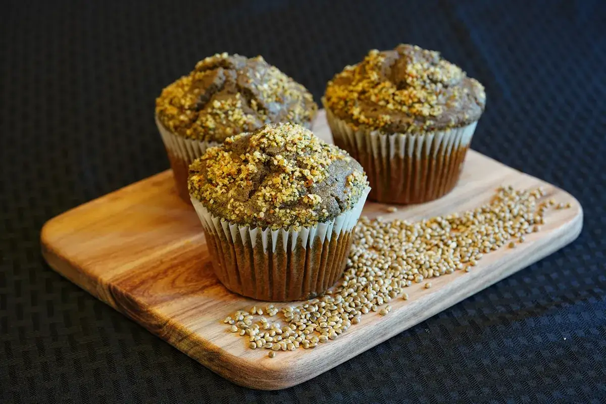 Drei Muffins mit Hanfsamen-Topping auf einem Holzbrett, daneben eine Streuung von Hanfsamen. Die Hanfsamen-Wirkung zeigt sich in diesen gesunden Leckereien.