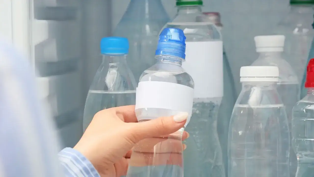 Dłoń trzyma butelkę z wodą, zastanawiając się, ile może stać woda w plastikowej butelce po otwarciu. W tle lodówka pełna innych butelek.