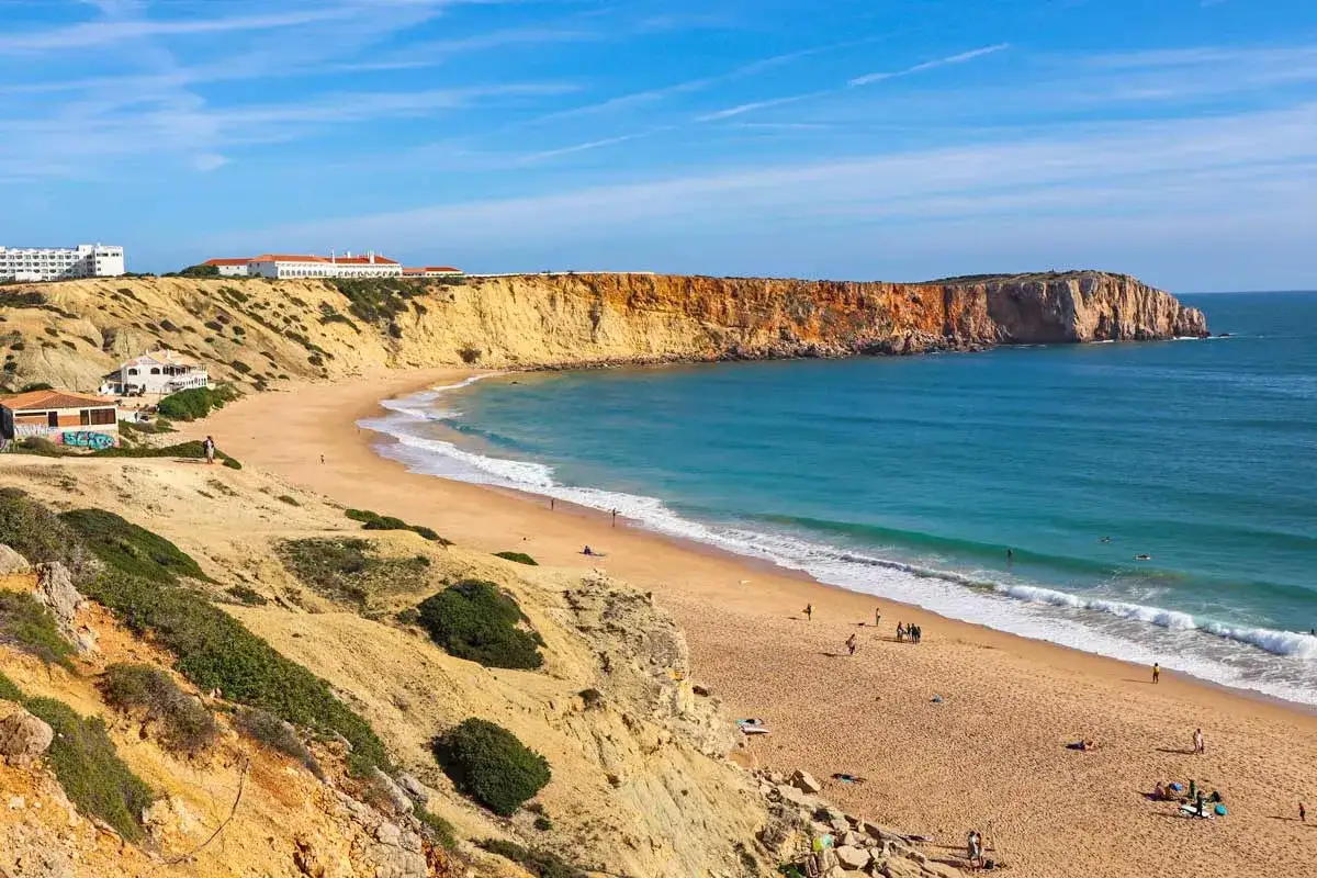Der **Sagres Strand** mit seinen goldenen Klippen und dem türkisfarbenen Meer lädt zum Entspannen ein. Einige Menschen genießen die Sonne, andere wagen sich ins Wasser.