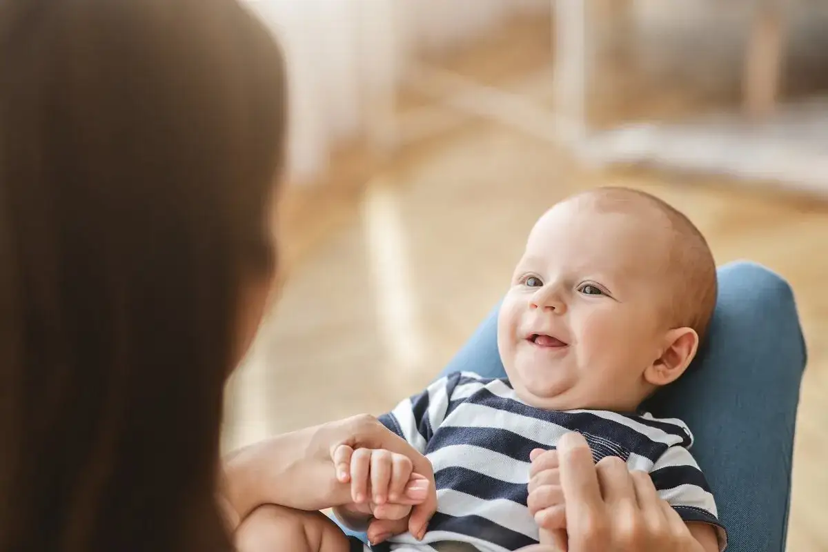 Uśmiechnięte miesięczne dziecko w pasiastej bluzeczce trzyma dłoń mamy.