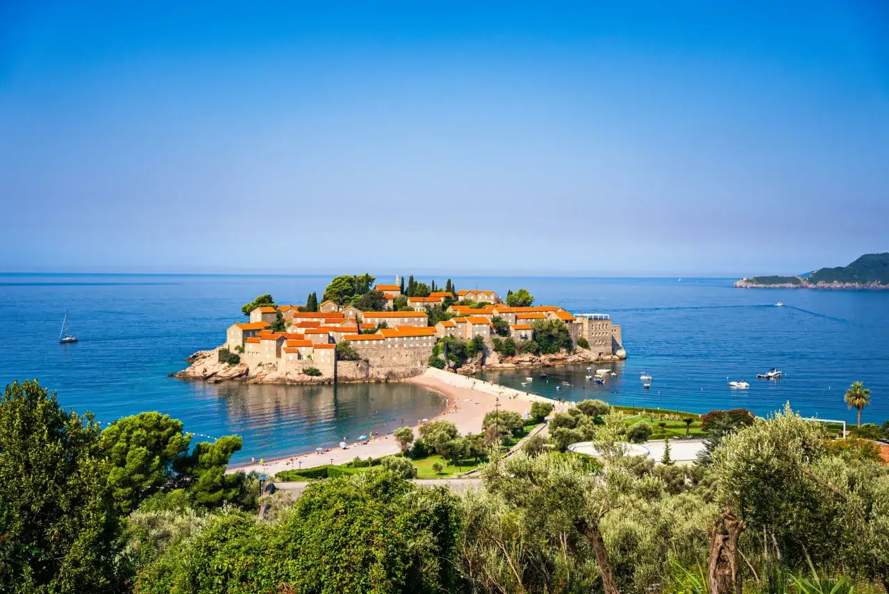 Słoneczna Czarnog&oacute;ra w czerwcu. Wyspa Sveti Stefan z kamiennymi domami i pomarańczowymi dachami, otoczona turkusowym morzem i piaszczystą plażą.