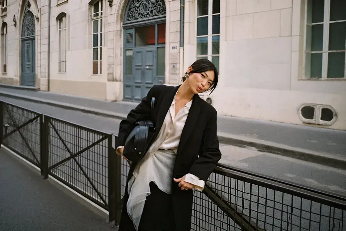 En kvinna i svart kavaj och vit blus bär en elegant svart väska. Hon lutar sig mot ett staket på en gata i Paris, med vackra byggnader i bakgrunden. Dessa söta väskor är perfekta för en stilfull dag ute.