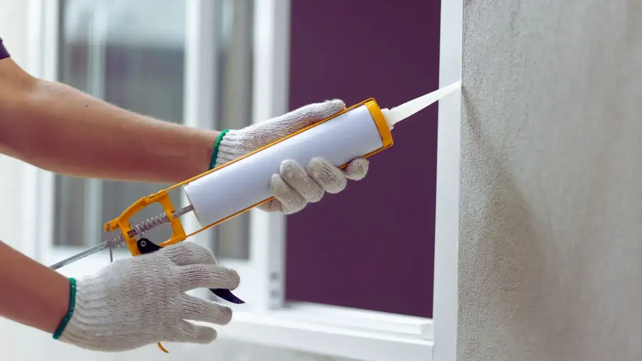 Mani guantate usano una pistola per silicone per sigillare un infisso. Il silicone serve a creare una barriera impermeabile e isolante.