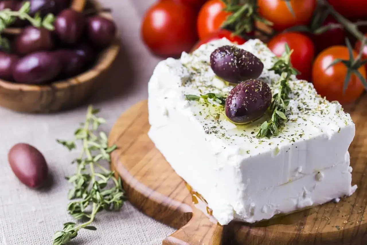 Un blocco di feta, formaggio di capra, condito con erbe e olive, su un tagliere con pomodorini e olive in sottofondo.