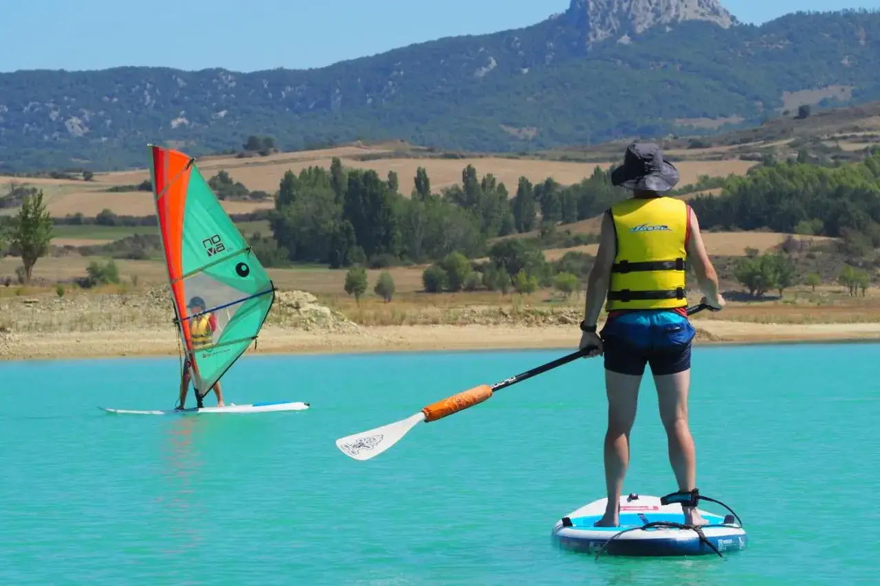 Un día soleado en el pantano de Alloz. Windsurf y paddle surf en aguas turquesas, con montañas verdes de fondo.
