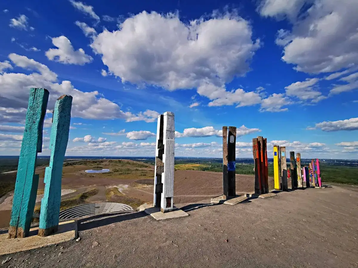 Bunte Holzskulpturen säumen den Weg auf der Halde Haniel. Der Himmel ist blau mit weißen Wolken.