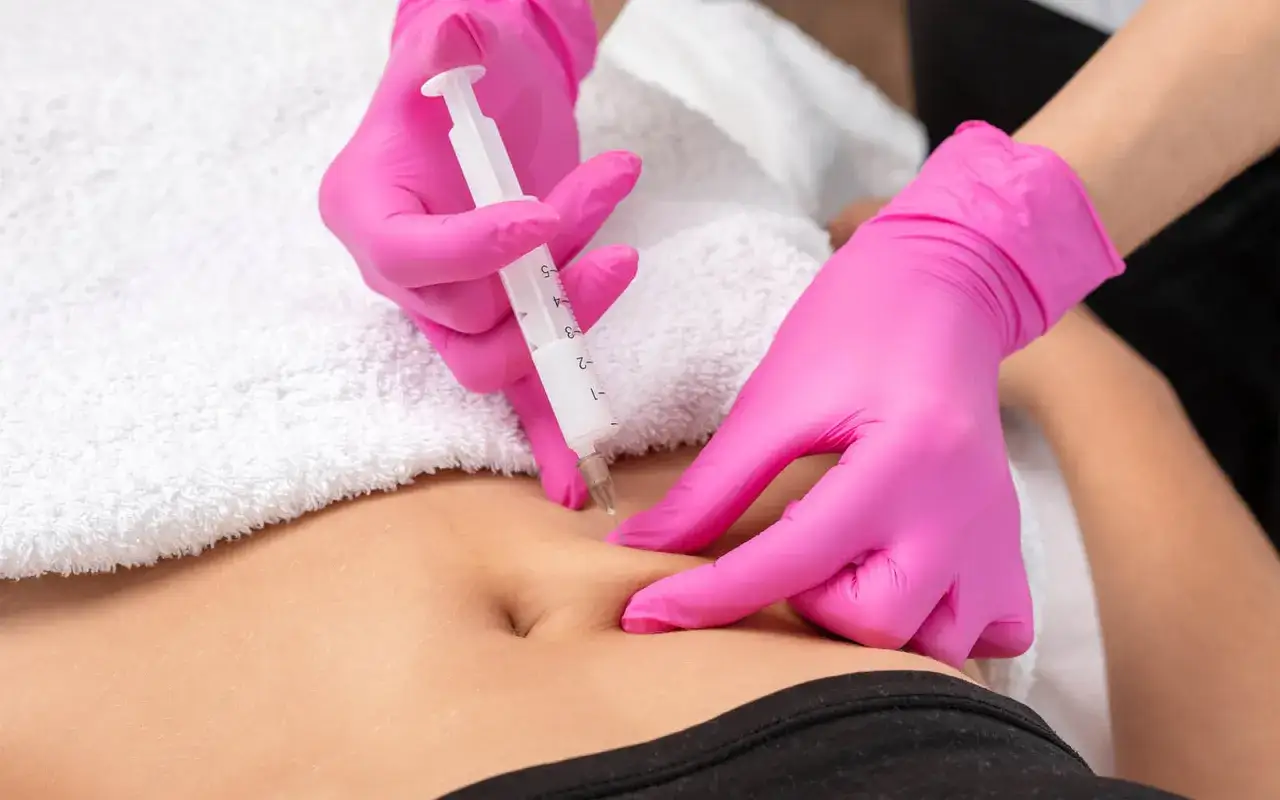 Hände in pinken Handschuhen führen eine Spritze im 90 Grad Winkel in die Bauchfalte einer Person.