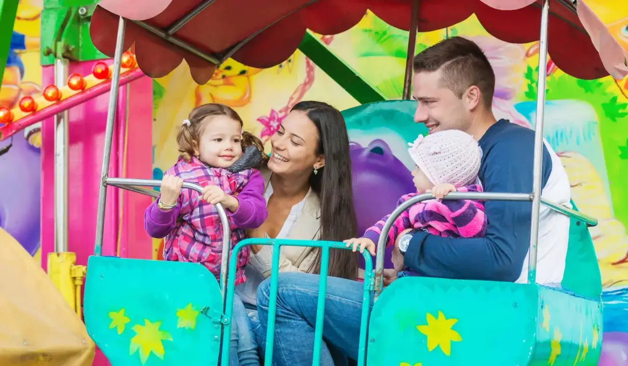 Familia feliz en un paseo de feria, uno de los muchos sitios para ir con niños. Las niñas ríen mientras los padres las observan.