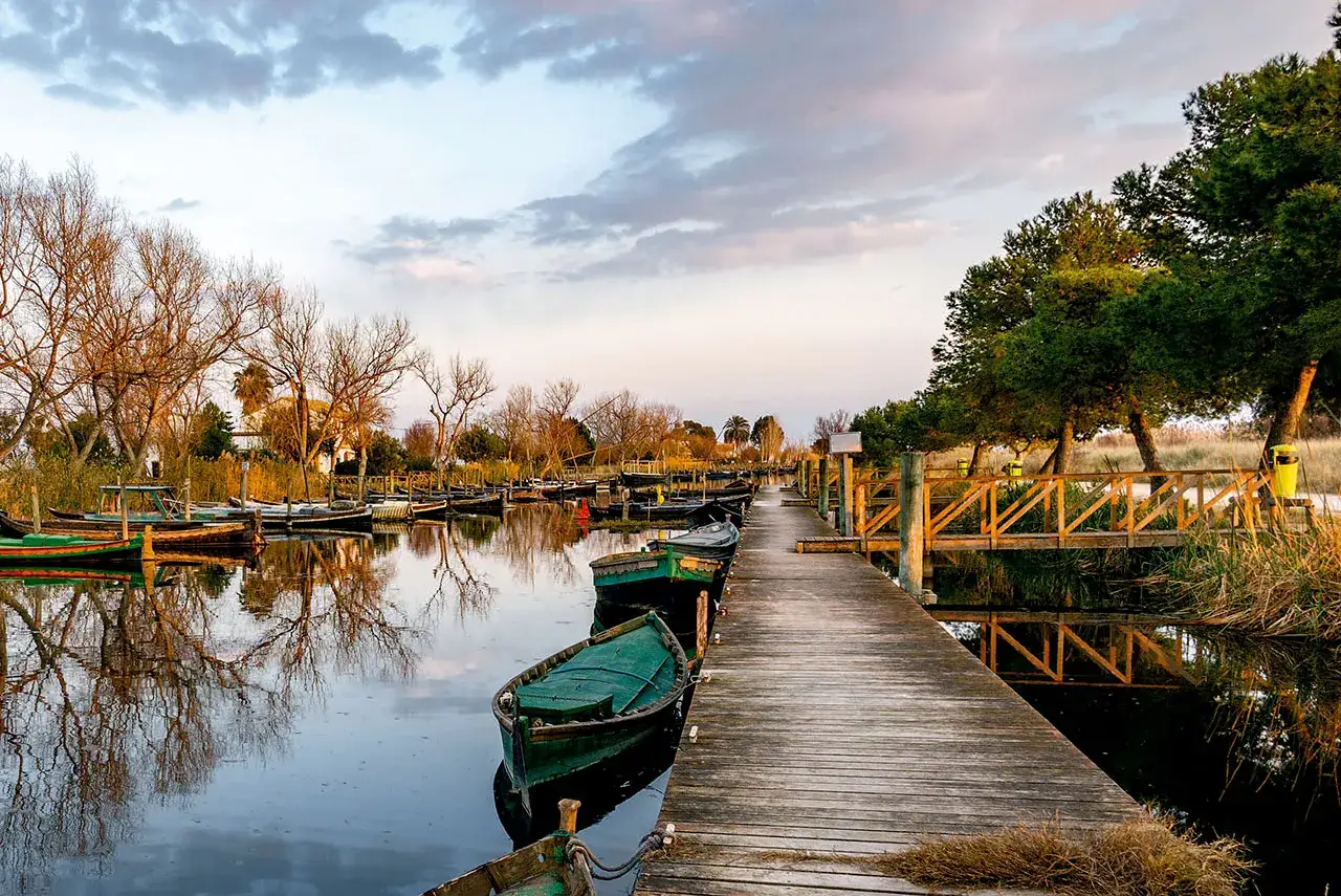 Embarcadero de madera con barcas verdes reflej&aacute;ndose en los lagos en Valencia. El atardecer ti&ntilde;e el cielo de tonos c&aacute;lidos.