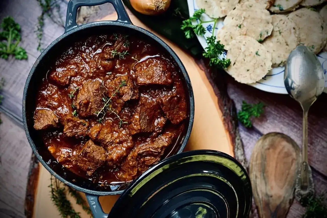 Herzhaftes Gulasch im Topf, daneben Kl&ouml;&szlig;e und frische Kr&auml;uter. Ein perfektes Gulasch Rezept f&uuml;r kalte Tage.