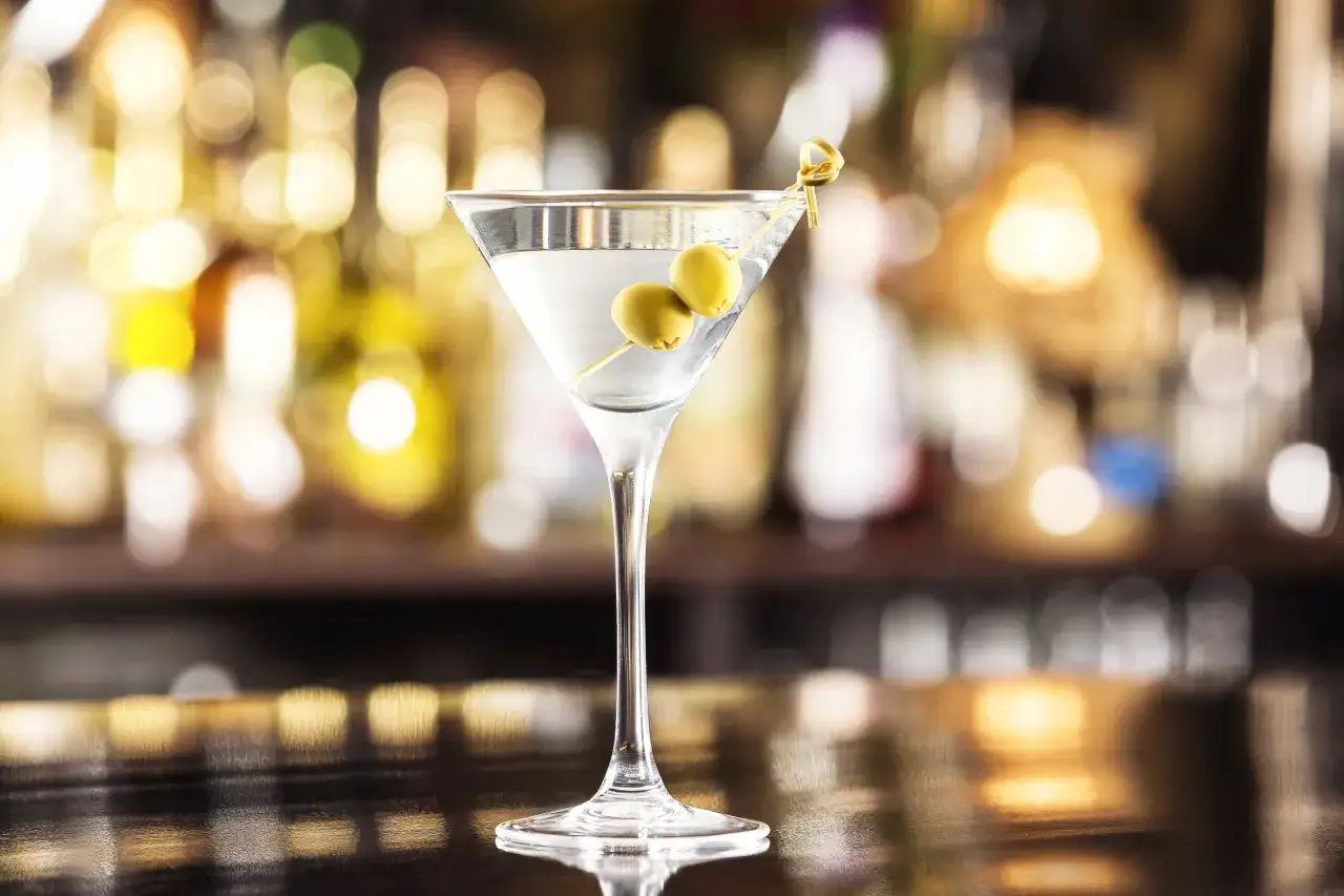 Klasyczny Martini wstrząśnięte czy zmieszane, ozdobione oliwkami, na tle rozmytych świateł baru.