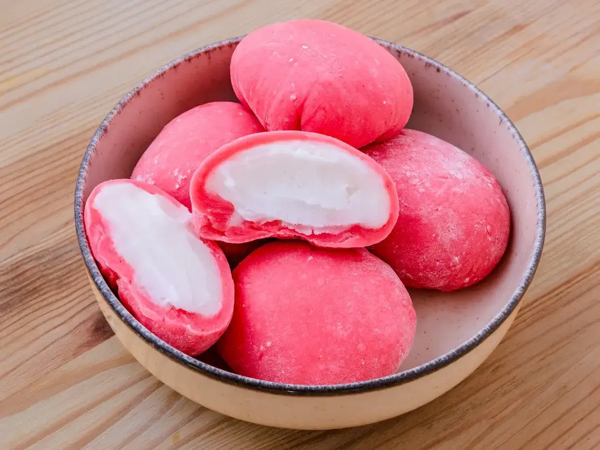 Un bol de mochis glacés roses, dont un coupé en deux révélant une garniture blanche crémeuse. Parfait pour une recette mochi glacé facile.
