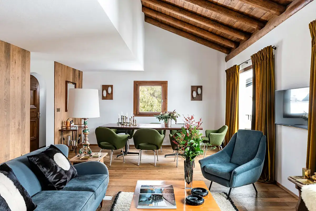 Soggiorno accogliente in una casa moderna con divano blu, poltrone verdi, tavolo da pranzo e travi a vista in legno.