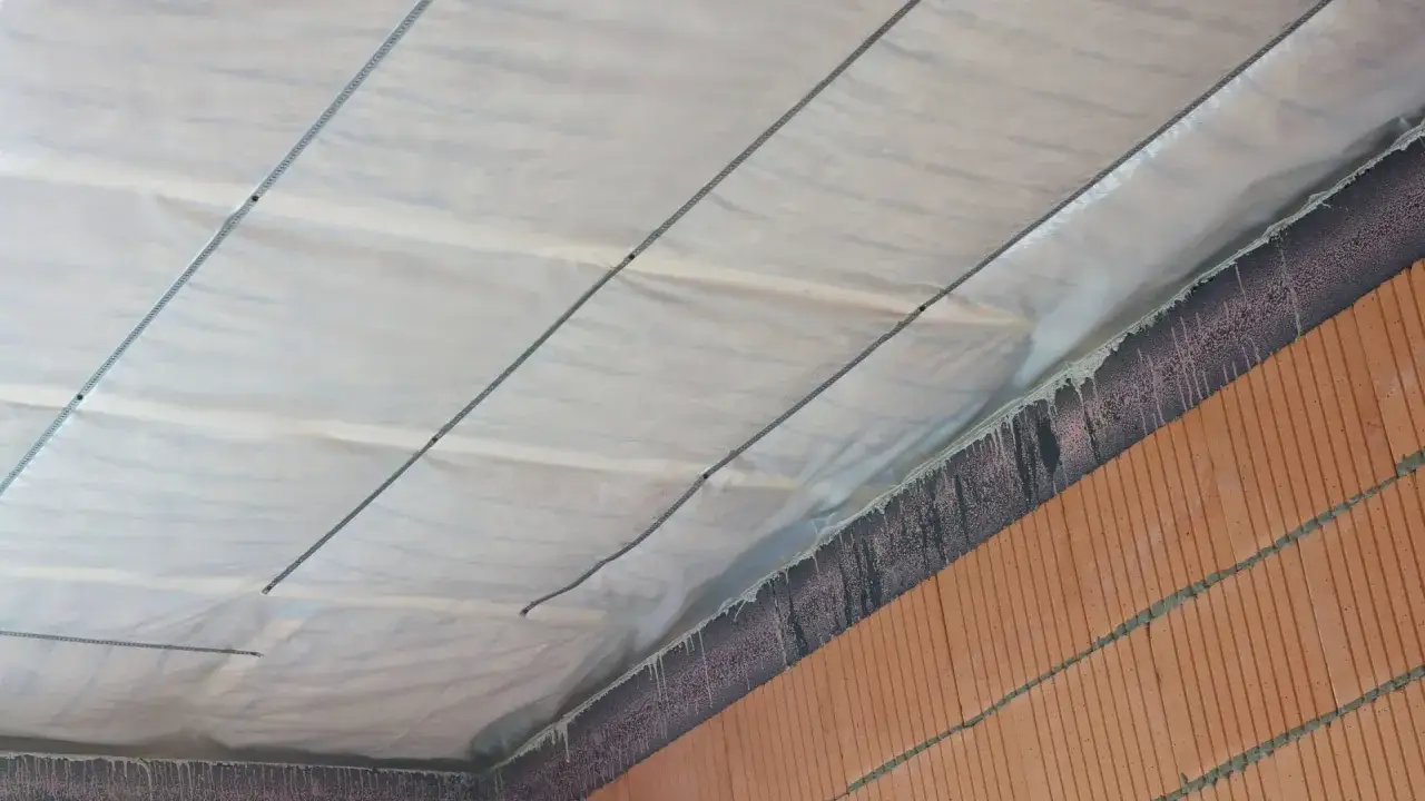 Die Kellerdecke wird mit einer wei&szlig;en Folie und Seilen isoliert. Die Ziegelwand darunter ist mit schwarzer Farbe bespr&uuml;ht.