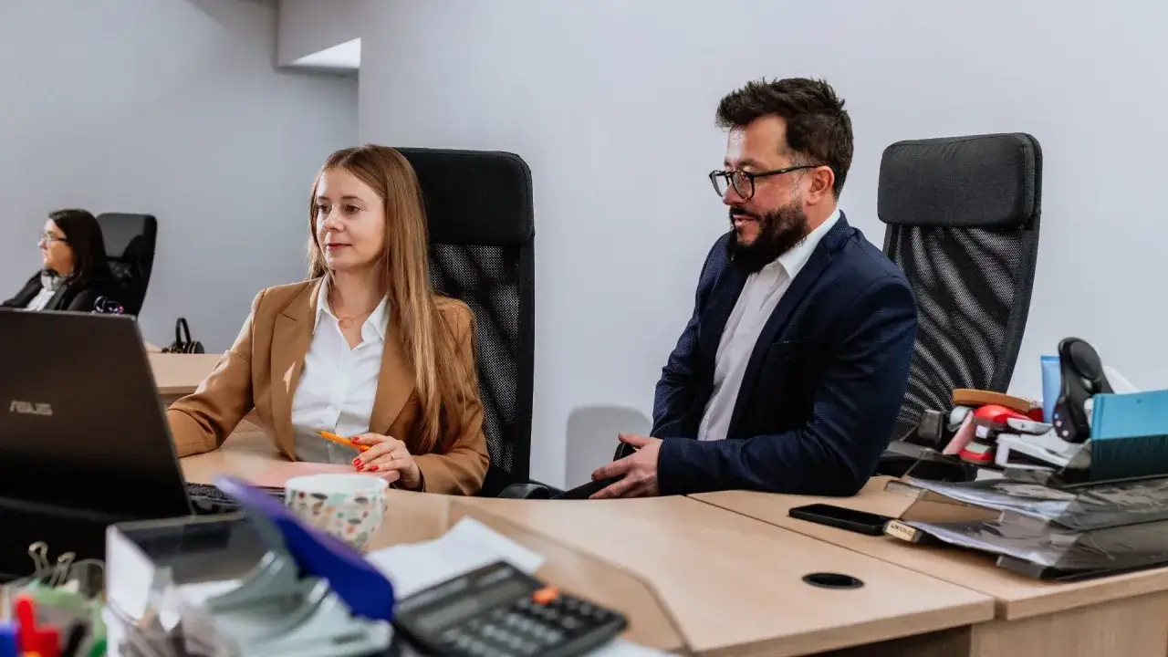 Kobieta i mężczyzna pracują przy biurku w biurze, na pierwszym planie laptop i kalkulator.