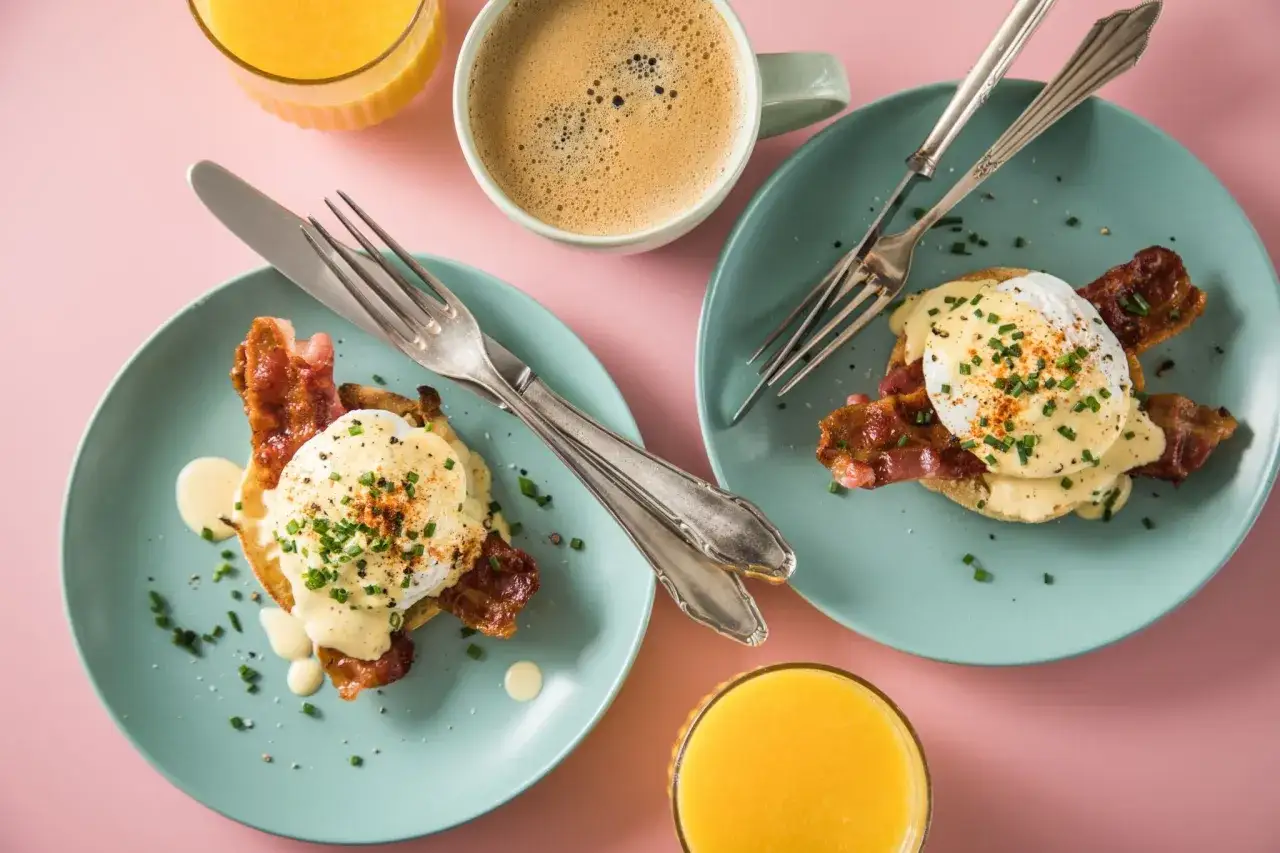 Zwei Teller mit Egg Benedict, dazu Kaffee und Orangensaft auf rosa Untergrund.