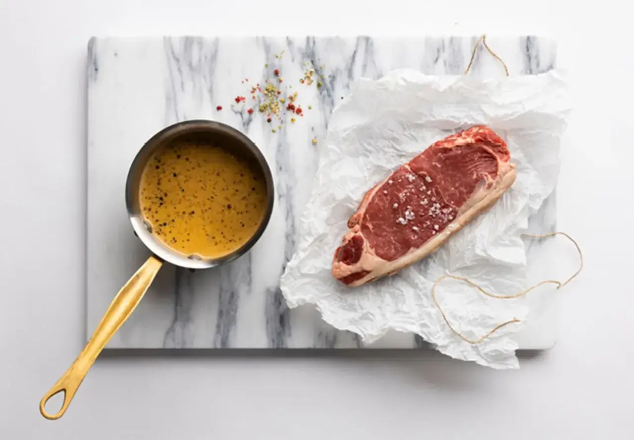 En r&aring; biff med salt och peppar ligger p&aring; sm&ouml;rpapper bredvid en kastrull med en gyllene peppars&aring;s. Recept p&aring; peppars&aring;s h&auml;r!