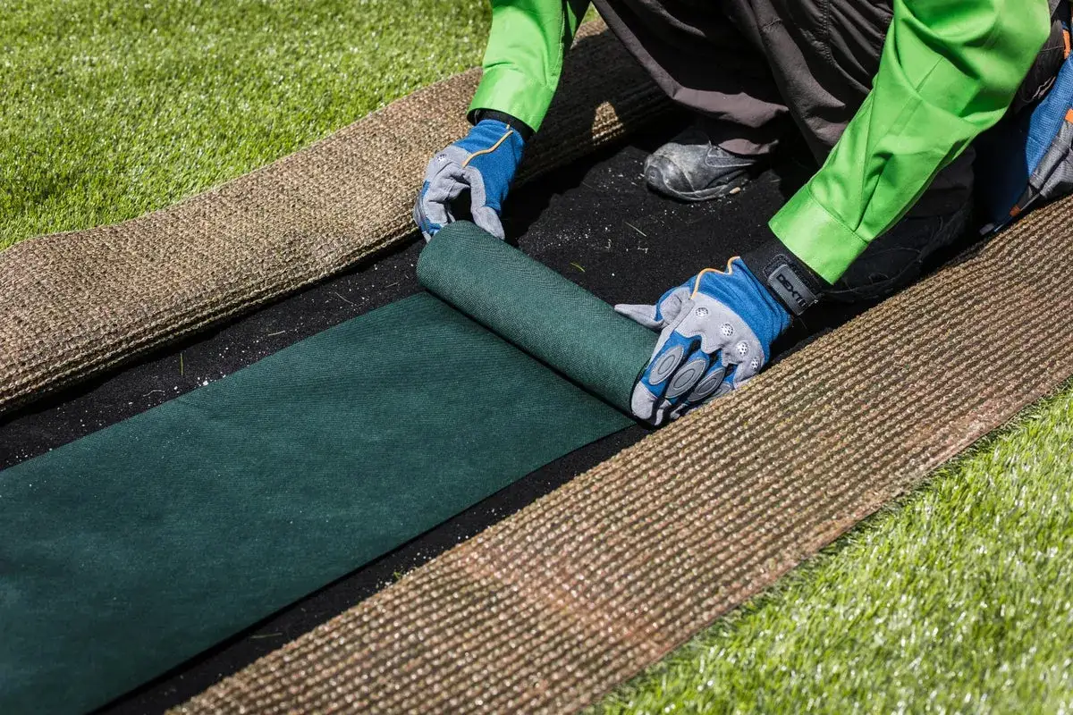 Manos enguantadas desenrollan c&eacute;sped artificial verde sobre una capa negra, prepar&aacute;ndose para poner c&eacute;sped artificial sobre c&eacute;sped natural.