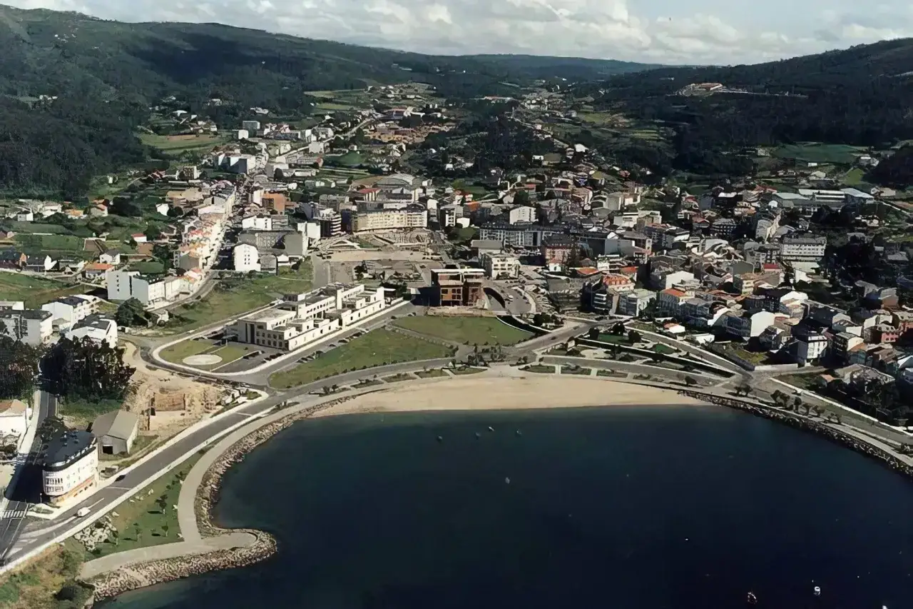Vista aérea de la playa de Cee, con el mar azul, la arena dorada y la ciudad extendiéndose hacia las colinas verdes.