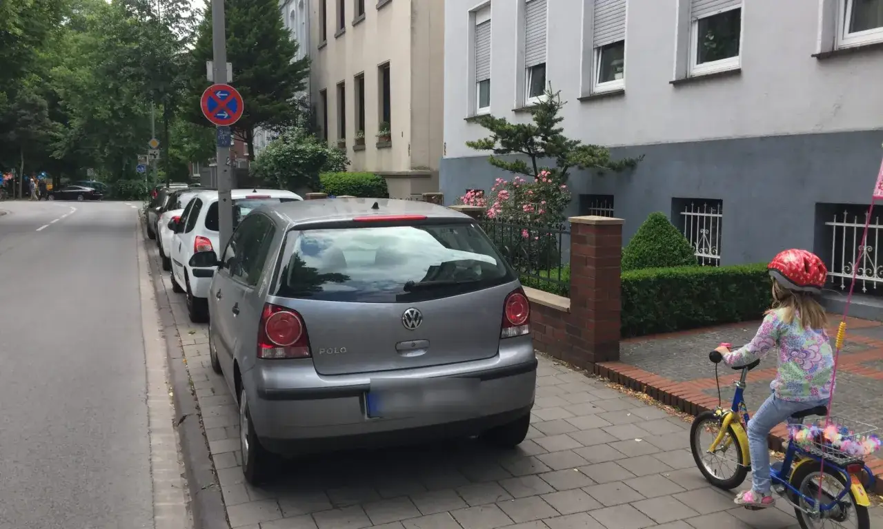 Verkehrsschild weist darauf hin: Fahrrad abstellen verboten. Autos parken am Straßenrand, ein Kind fährt Fahrrad.