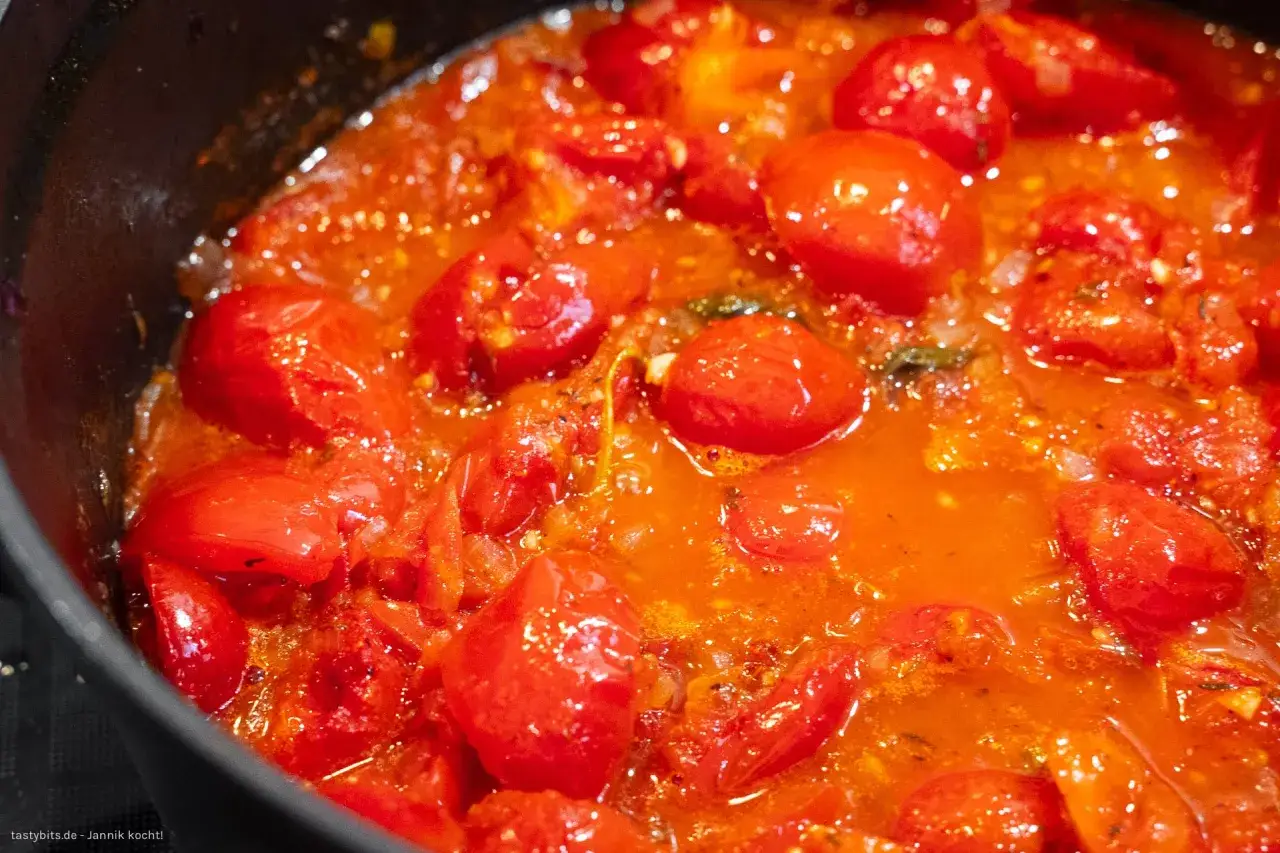 Ein köstliches, blubberndes Tomaten Sugo in einer schwarzen Pfanne, voller saftiger Kirschtomaten und aromatischer Kräuter.