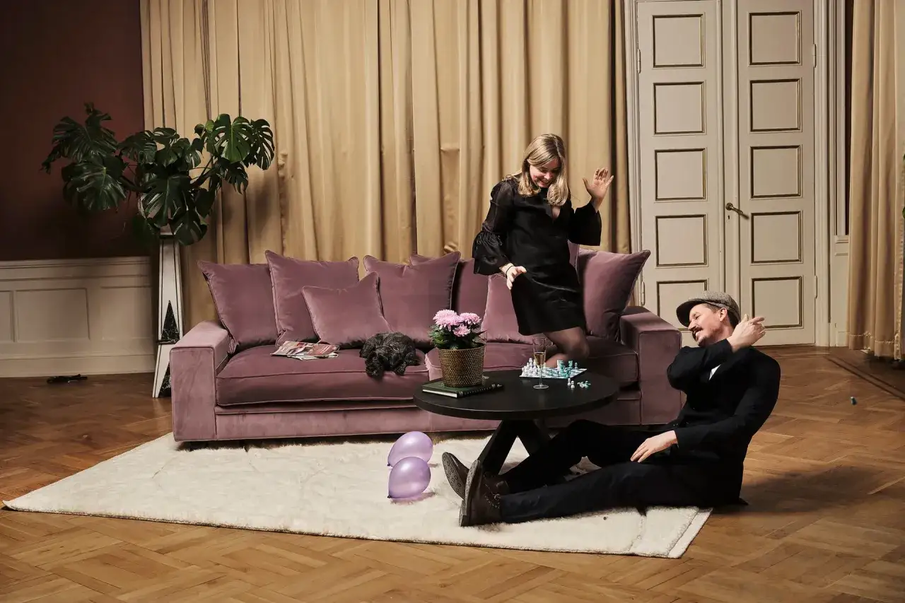 En kvinna dansar p&aring; en dyr soffa medan en man sitter p&aring; golvet. En hund vilar p&aring; soffan.
