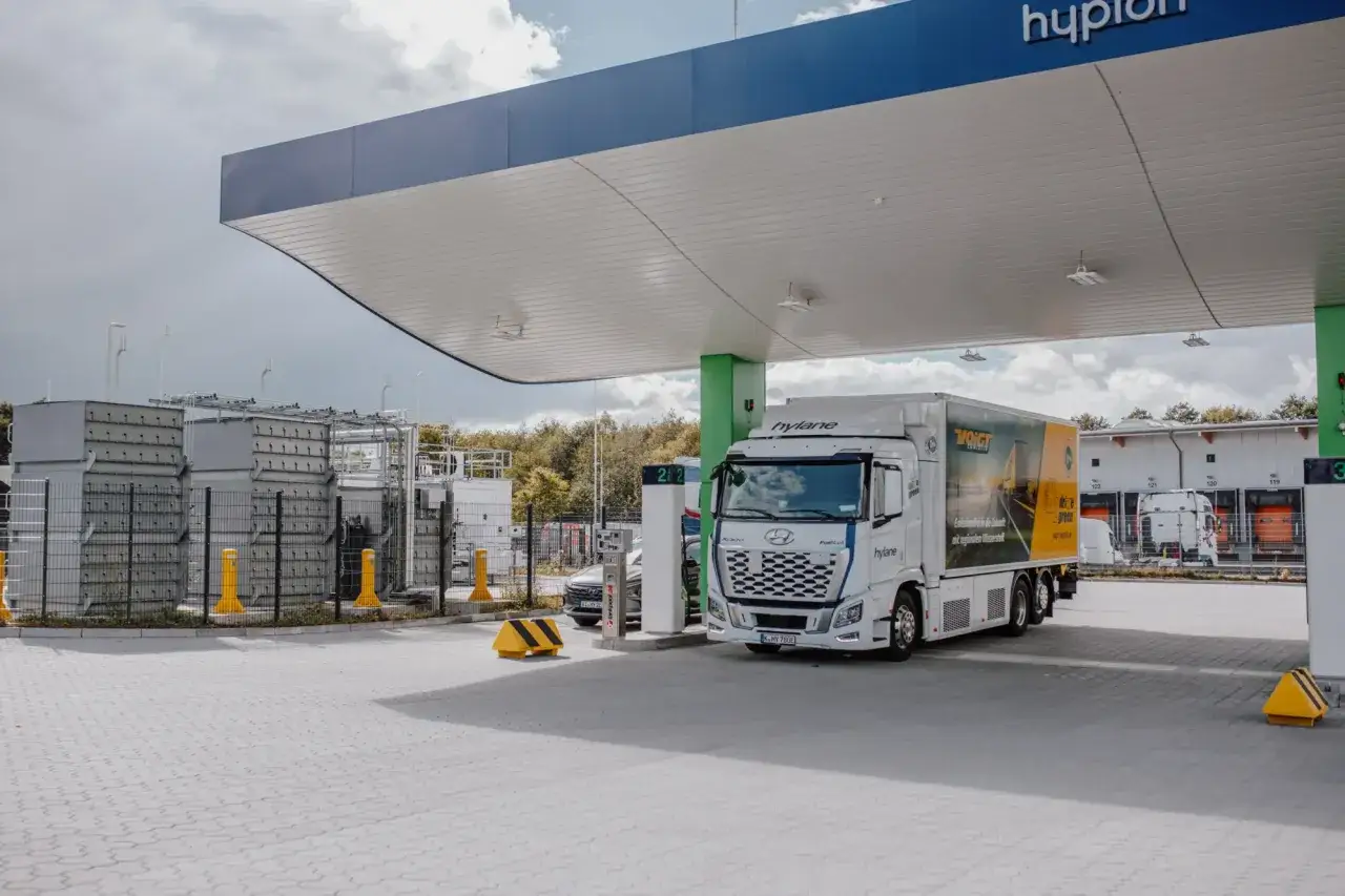 Cami&oacute;n de hidr&oacute;geno Hylane en una estaci&oacute;n de servicio. El veh&iacute;culo blanco con remolque amarillo y negro se detiene bajo un techo azul y blanco.
