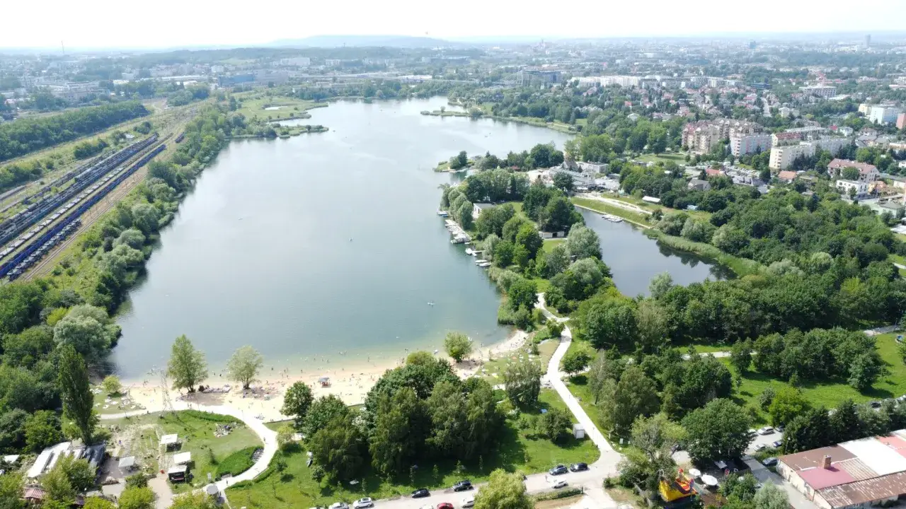 Plaża Bagry tętni życiem, ludzie odpoczywają nad wodą, a w oddali widać miasto i tory kolejowe.