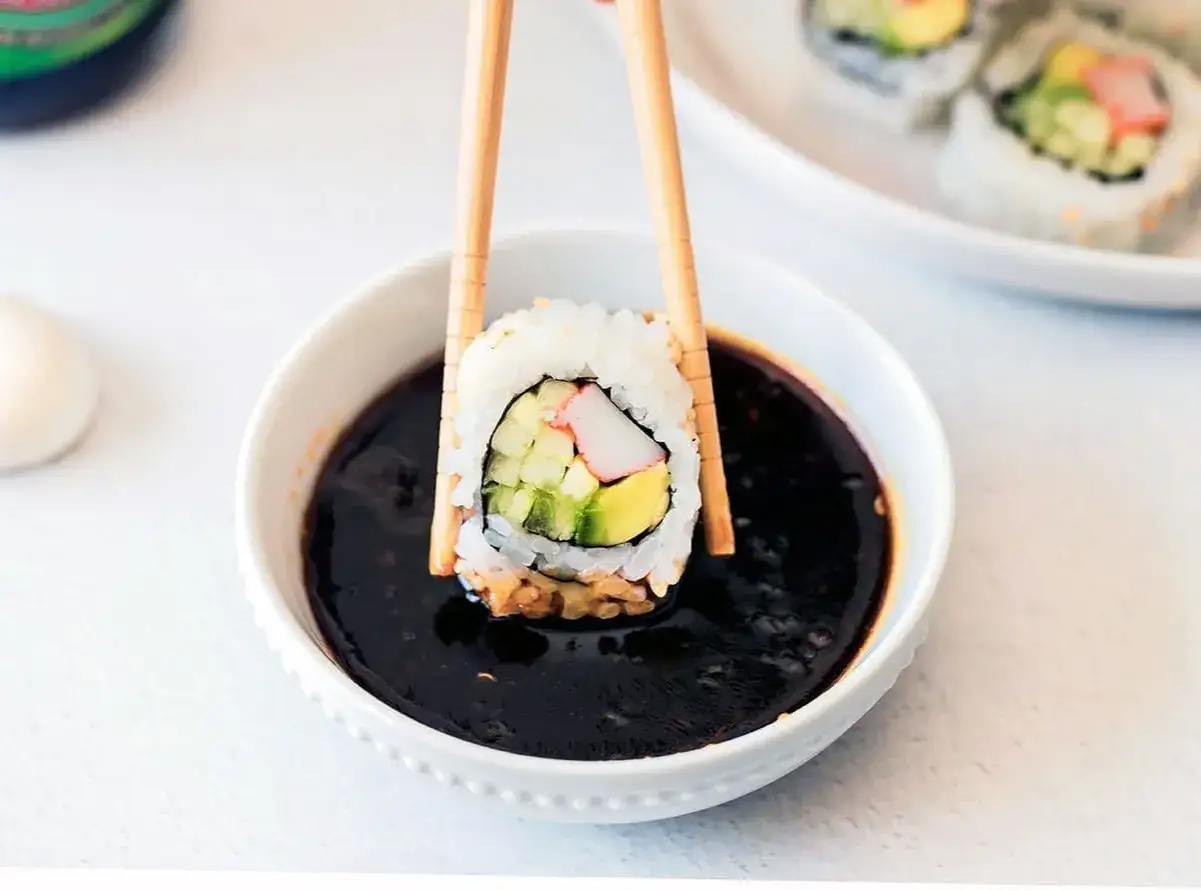 Pałeczkami zanurzam sushi w gęstym słodkim sosie do sushi. Widać kawałki awokado, ogórka i paluszka krabowego.