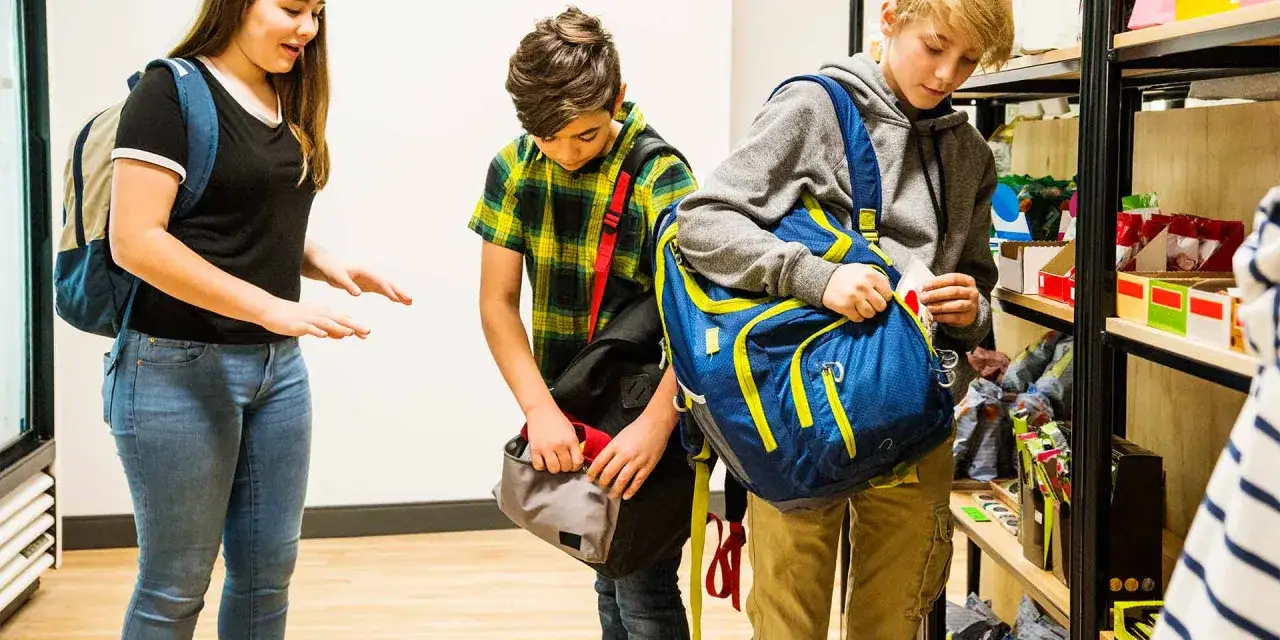 Drei Kinder mit Rucks&auml;cken in einem Gesch&auml;ft. Eines steckt etwas in seine Tasche, was auf Kleptomanie bei Kindern hindeuten k&ouml;nnte.