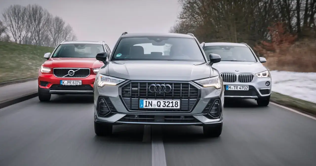Szary Audi Q3 2025 na pierwszym planie, z czerwonym Volvo i srebrnym BMW w tle, jadące drogą.