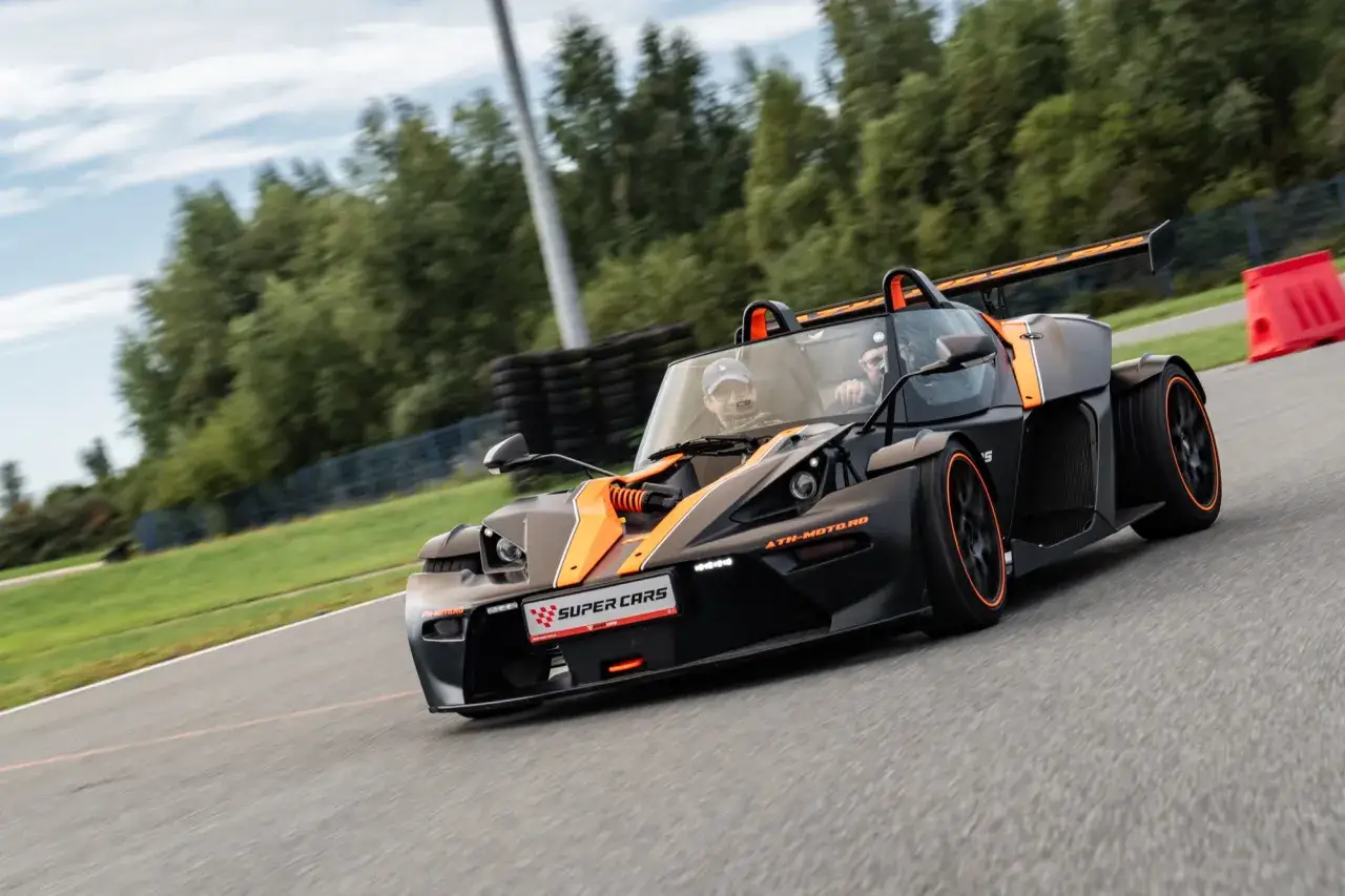 Czarny KTM X-Bow GT XR z pomarańczowymi akcentami pędzi po torze wyścigowym, kierowca w czapce z daszkiem i pasażer cieszą się jazdą.