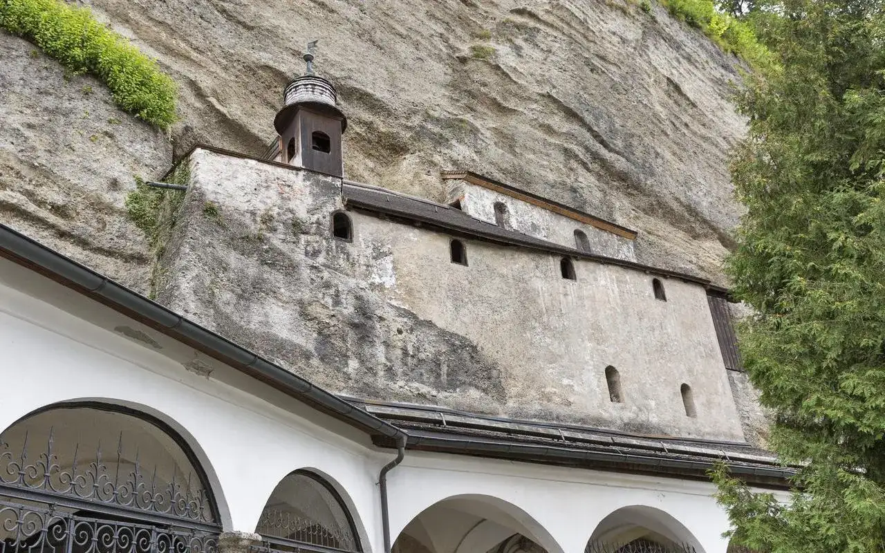 Teil der Erzabtei Salzburg, ein Geb&auml;ude schmiegt sich an eine Felswand, mit einem kleinen Turm und Arkaden im Vordergrund.