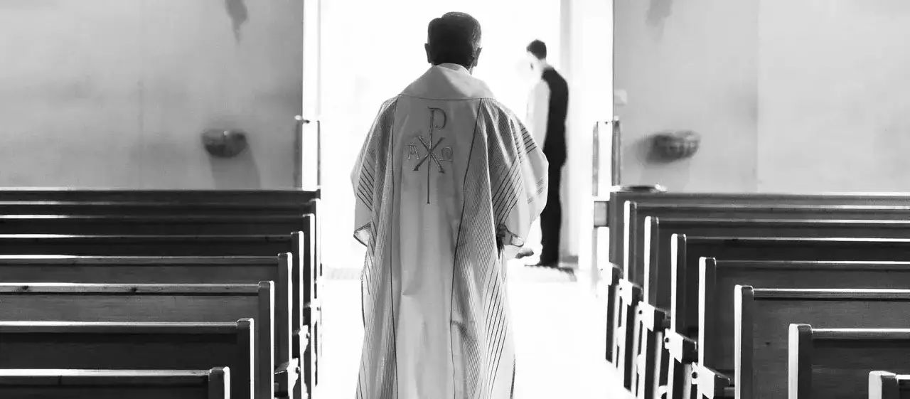 Ein katholischer Pfarrer im Messgewand mit dem Chi-Rho-Symbol geht durch Kirchenb&auml;nke. Die Frage, ob ein katholischer Pfarrer heiraten darf, besch&auml;ftigt viele.