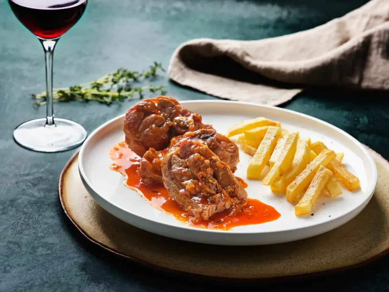 Delicioso cordero al chilindrón en un plato blanco, acompañado de patatas fritas y una copa de vino tinto.