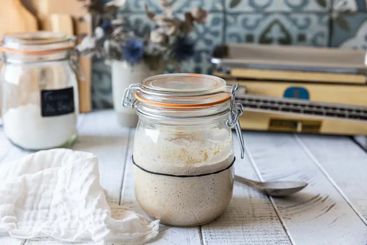 Un bocal en verre rempli de levain rafraichi, pr&ecirc;t pour la prochaine fourn&eacute;e de pain. Une cuill&egrave;re en bois et un torchon sont &agrave; proximit&eacute;.