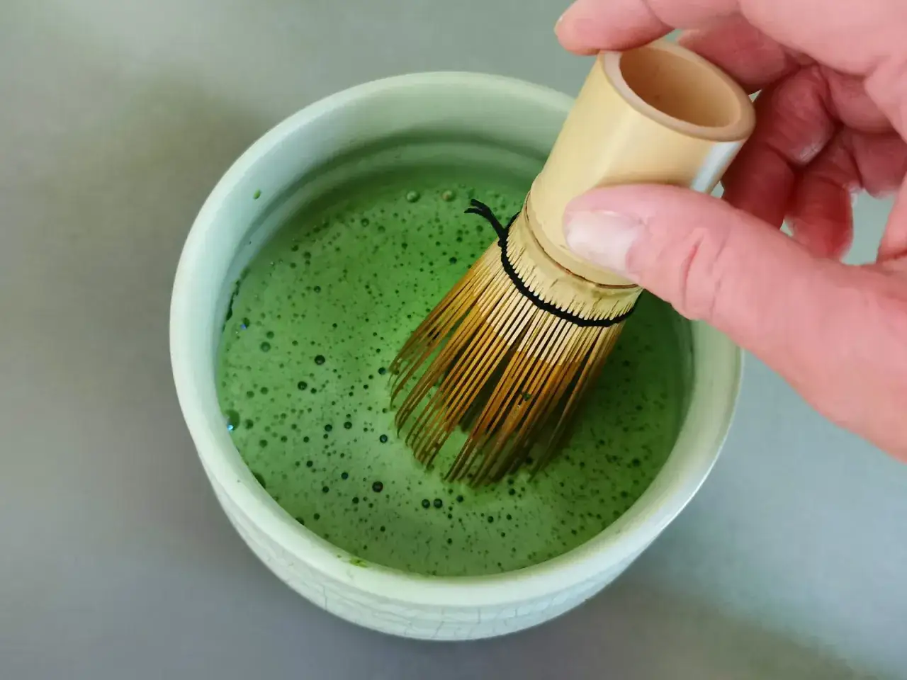 Hand whisking bright green matcha powder in a bowl, creating a frothy, slightly bitter drink.