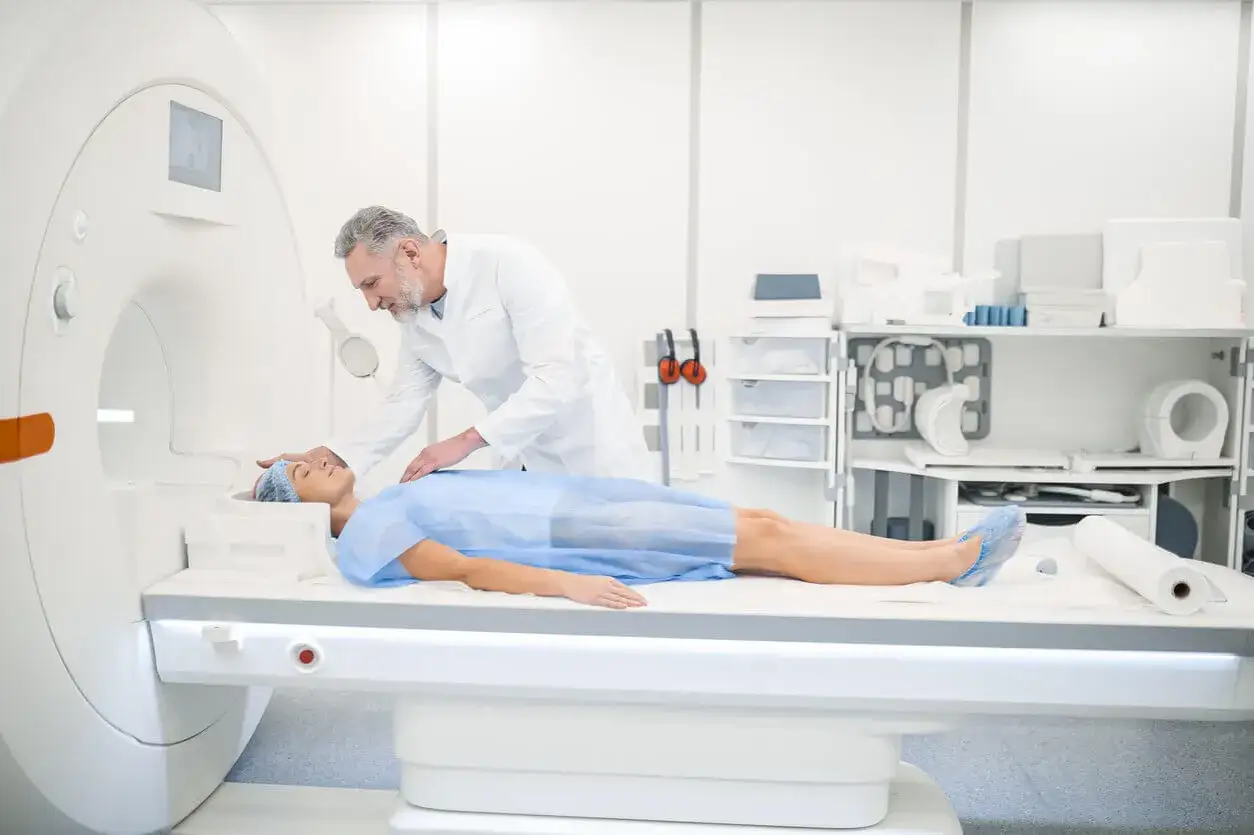 Pacjentka w fartuchu medycznym leży na stole tomografu. Lekarz przygotowuje ją do badania. Czy do tomografii trzeba się rozbierać? Zazwyczaj tak, ale w tym przypadku pacjentka ma na sobie fartuch.