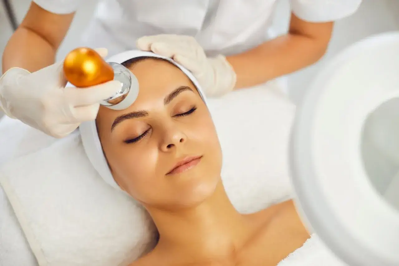 Tratamiento facial para combatir la flacidez facial a los 40. Una esteticista usa un dispositivo dorado para rejuvenecer la piel.