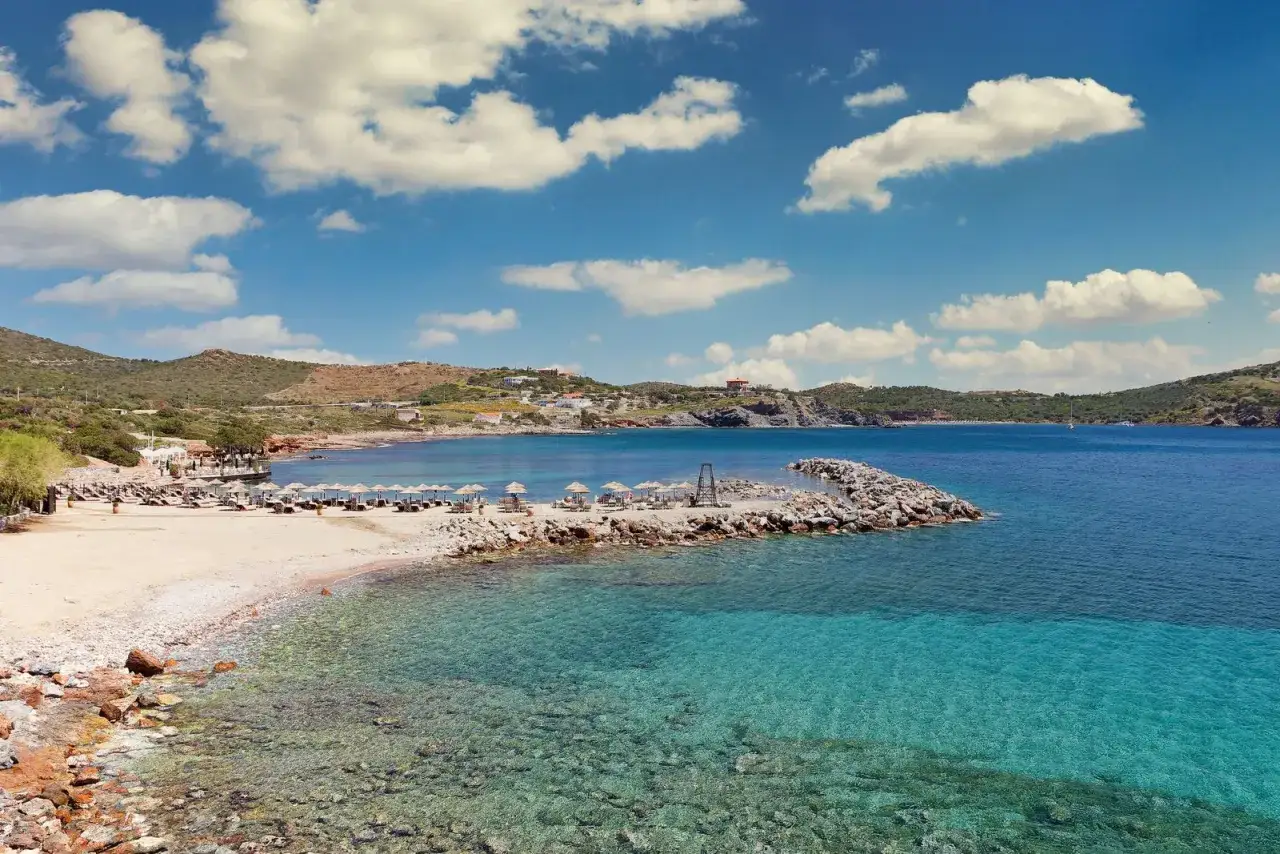 Piękna plaża w Atenach z turkusową wodą, leżakami i parasolami. W oddali widać wzgórza i zabudowania.