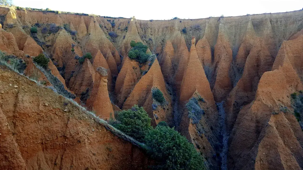 Paisaje erosionado con formaciones c&oacute;nicas de tierra rojiza, como un pont&oacute;n de la oliva, salpicado de vegetaci&oacute;n verde.